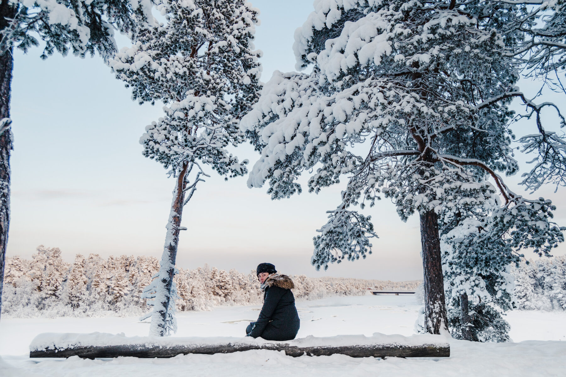 Mulher em um tronco coberto de neve de frente para lago congelado Inari, finlândia