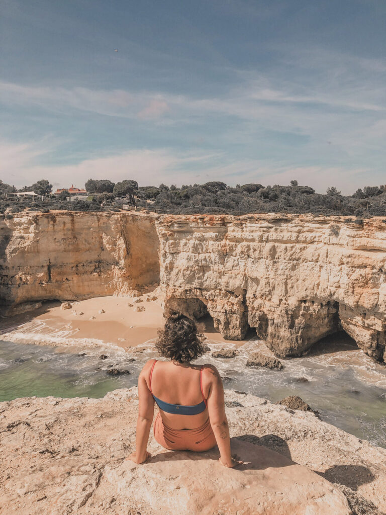 mulher sentada em cima do arco de albandeira de costas para a camera observando a vista das falésias e do mar no algarve em portugal