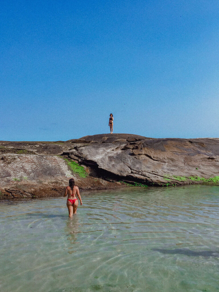 duas mulheres na praia do secreto, uma mulher de biquini preto no topo da pedra e uma mulher de biquini vermelho andando na água em direção a pedra