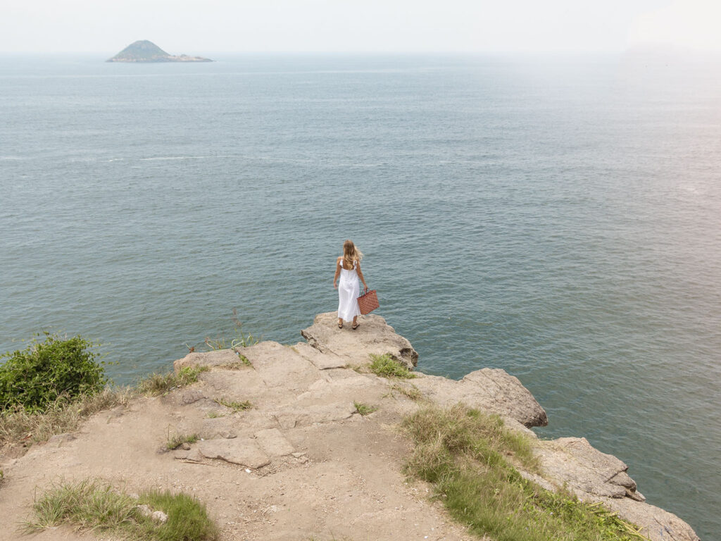 Mulher loira segurando bolsa de vestido branco na ponta da vista da praia da joatinga